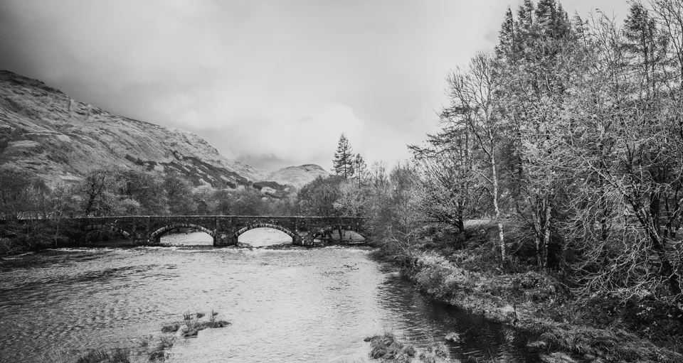 A stone bridge crossing a river beneath wooded hills and clouds, photographed in black and white.